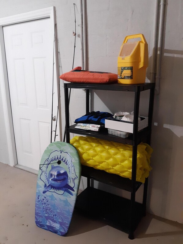 Une étagère en métal noir sur laquelle se trouvent un récipient jaune, un coussin rouge et une planche de surf bleue et jaune appuyée contre un mur.