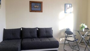 Un salon avec un canapé d’angle gris, une petite table ronde avec des chaises et deux tableaux encadrés accrochés au mur.
