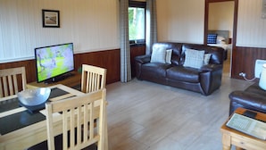Un salon doté d’un plancher en bois, d’un canapé en cuir marron, d’une table à manger en bois avec des chaises, d’un téléviseur et d’une fenêtre avec des rideaux.