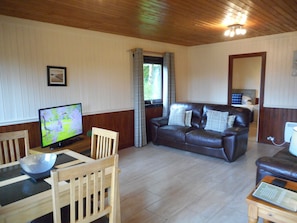 Een woonkamer met een houten vloer, een bruine leren bank, een houten eettafel met stoelen, een televisie en een raam met gordijnen.
