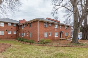 A multi-story brick building with a flat roof, multiple windows, and a small front yard with a tree and shrubs.
