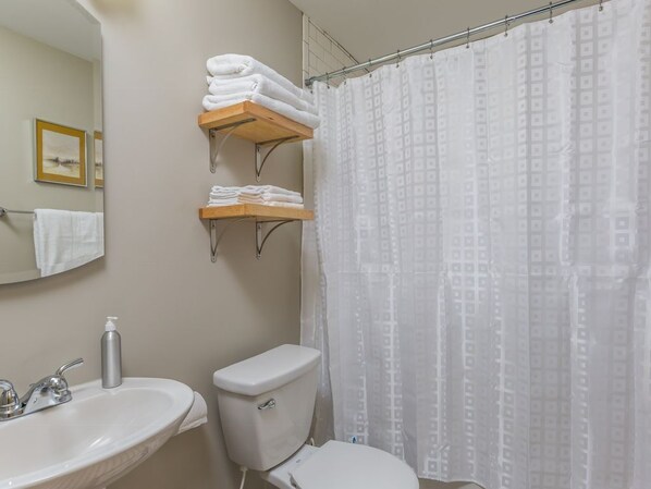 A bathroom with a white sink, toilet, and shower curtain. There are two wooden shelves above the toilet holding folded towels.