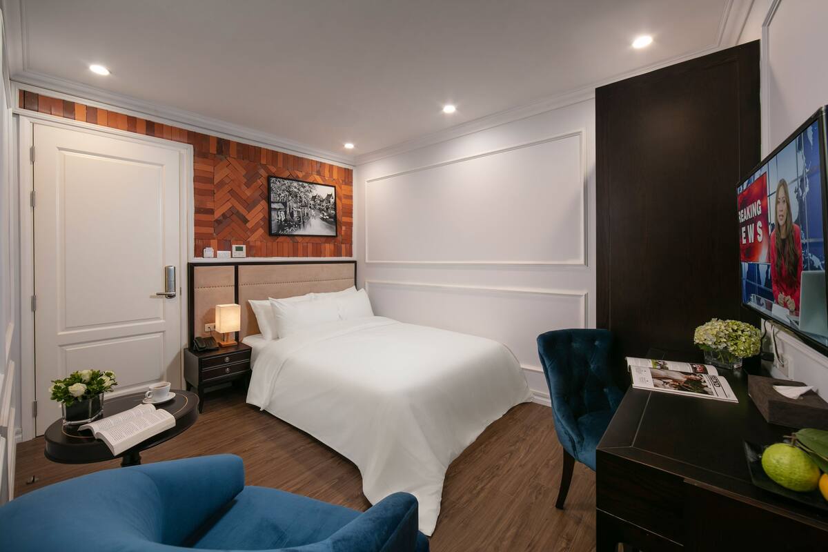 Ein Hotelzimmer mit Bett, Schreibtisch, Stuhl, Nachttisch und einem an der Wand montierten Fernseher.