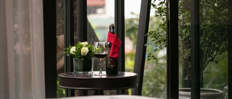 A table with a bottle of wine, a glass, and a vase with flowers, set against a window with a view of greenery.