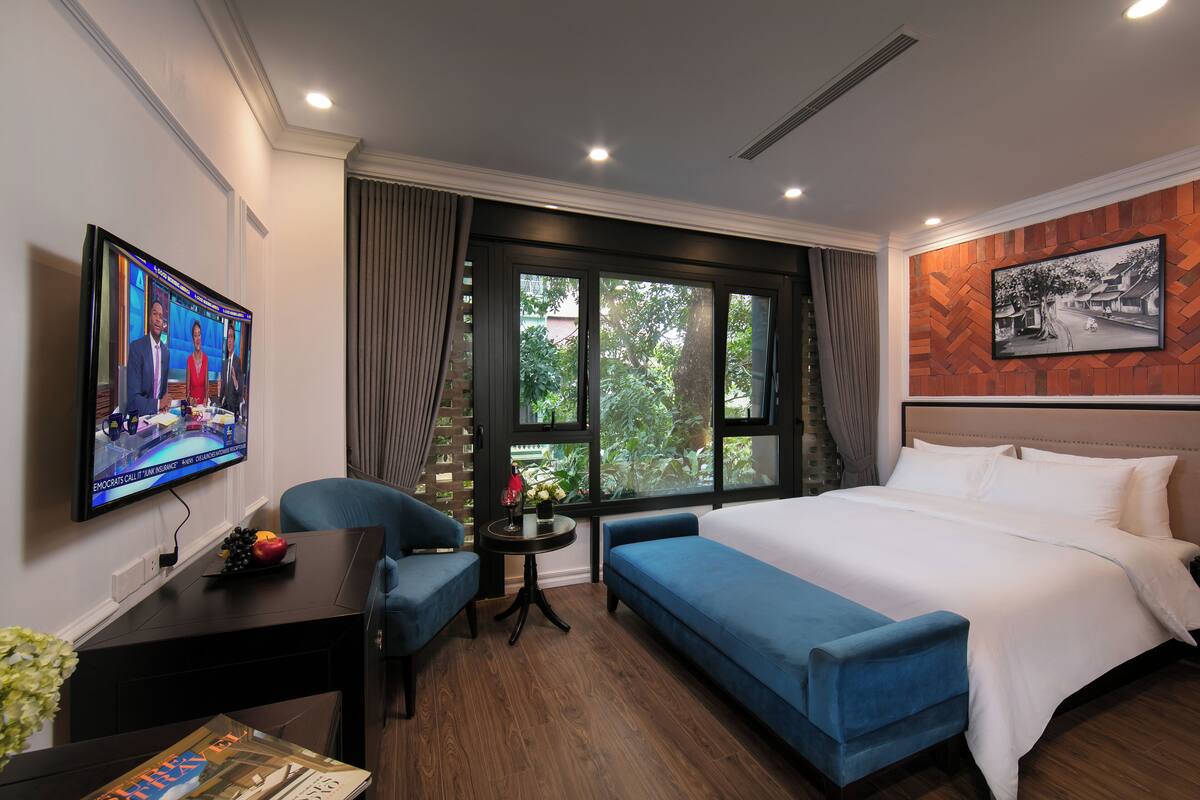 Ein Hotelzimmer mit einem großen Bett, einem blauen Sofa, einem kleinen Tisch mit einer Vase, einem an der Wand montierten Fernseher und einem Fenster mit Blick auf Grünflächen.