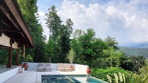 Une piscine dotée de chaises longues et d’une pergola, entourée d’une végétation luxuriante et offrant une vue sur un paysage montagneux.