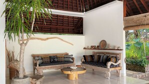 A spacious indoor area with a high ceiling, two wooden sofas with patterned cushions, a large potted plant, and a stone wall. - Villa Sari Lombok, Hidden treasure (Lombok)