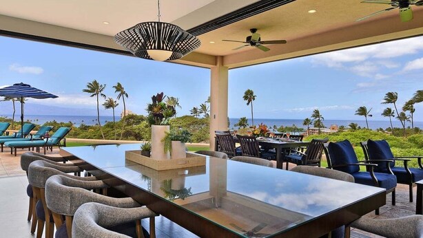 A spacious outdoor dining area with a glass table and chairs, overlooking a scenic view of the ocean and palm trees.