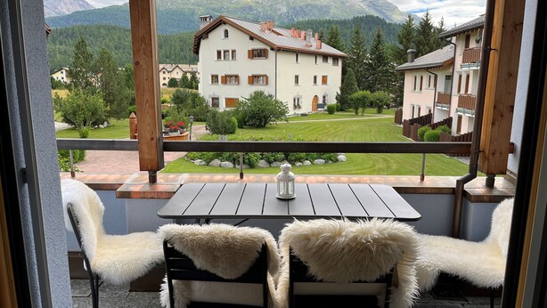 Un balcone con due sedie e un tavolo, affacciato su un paesaggio montano con edifici.