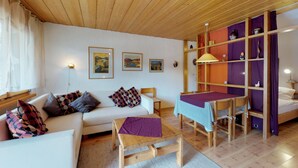 Een gezellige woonkamer met een witte bank, een houten salontafel en een eettafel met stoelen. Er hangen ingelijste foto’s aan de muur en een boekenkast met kleurrijke planken.