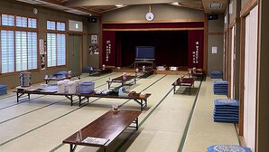 Une pièce de style japonais traditionnel, avec des tatamis, de basses tables et une scène surmontée d’un rideau rouge.