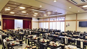 Une salle de conférence avec des tables dressées pour un repas, une scène avec des rideaux et un écran de télévision au mur.
