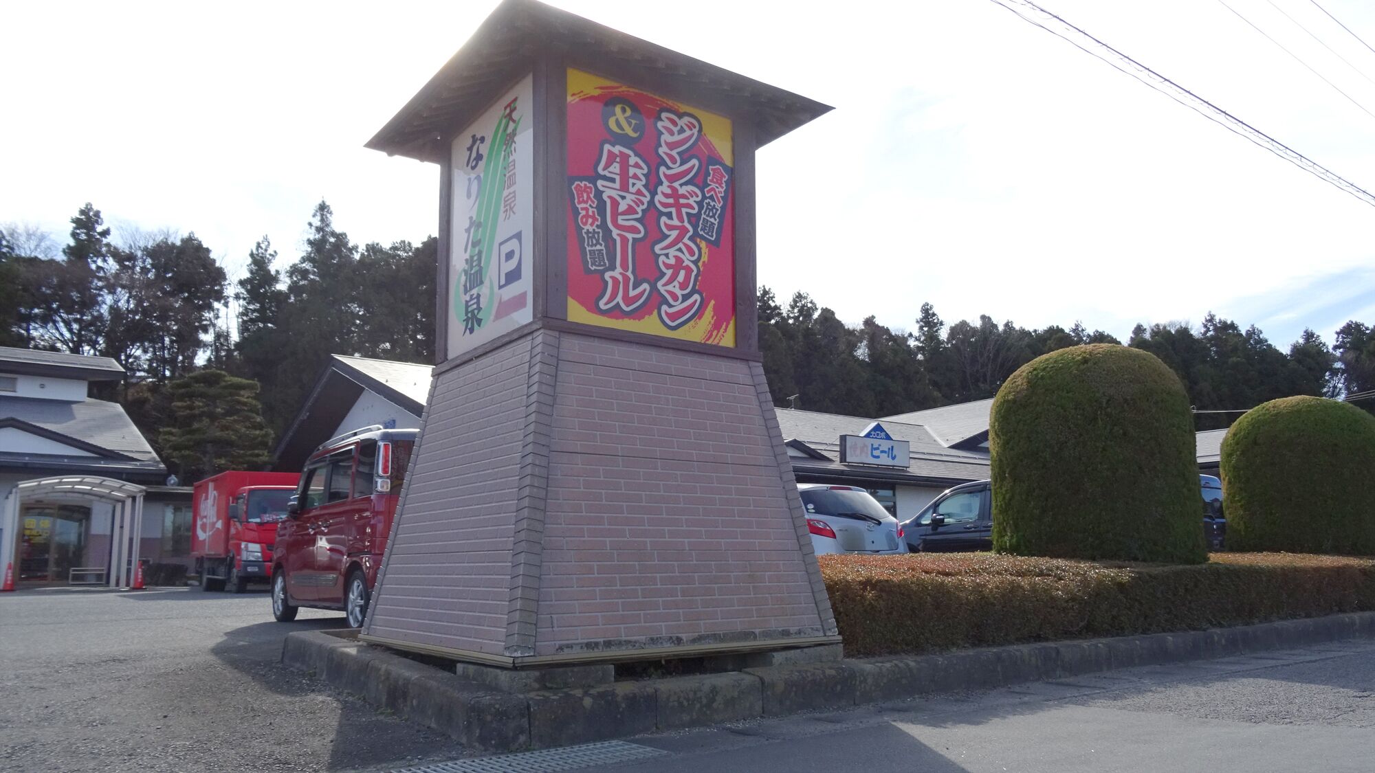 Un bâtiment avec une enseigne en japonais, un camion rouge garé devant, et une haie sur la droite.