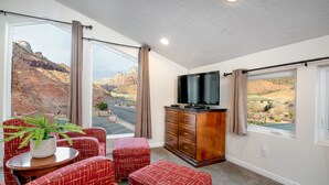 A room with a large window offering a view of a mountainous landscape, a flat-screen TV on a wooden cabinet, and red patterned furniture. - Apartment #1 GREAT LOCATION Zion National Park entrance as close as you can get! (Springdale)