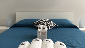A bed with a blue bedspread, a white headboard, and a black and white patterned pillow. There are four rolled white towels with a gray center placed in front of the bed. - H.ö.H (Porto)