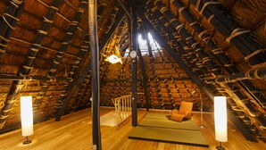 A cozy attic room with a thatched roof, wooden beams, a cushioned bench, and two standing lamps.