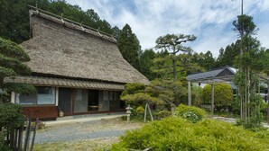 A traditional Japanese house with a thatched roof, surrounded by lush greenery and a gravel path.