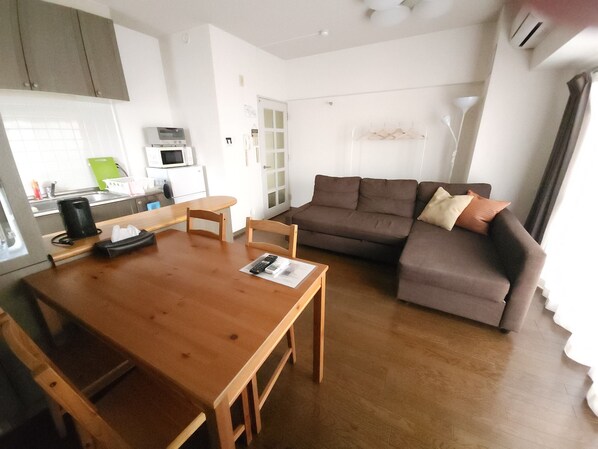 A living room with a wooden table, a brown sofa, and a kitchen area with a microwave and cabinets. - Convenient to Dotonbori,OsakaCastle (Ōsaka-shi)