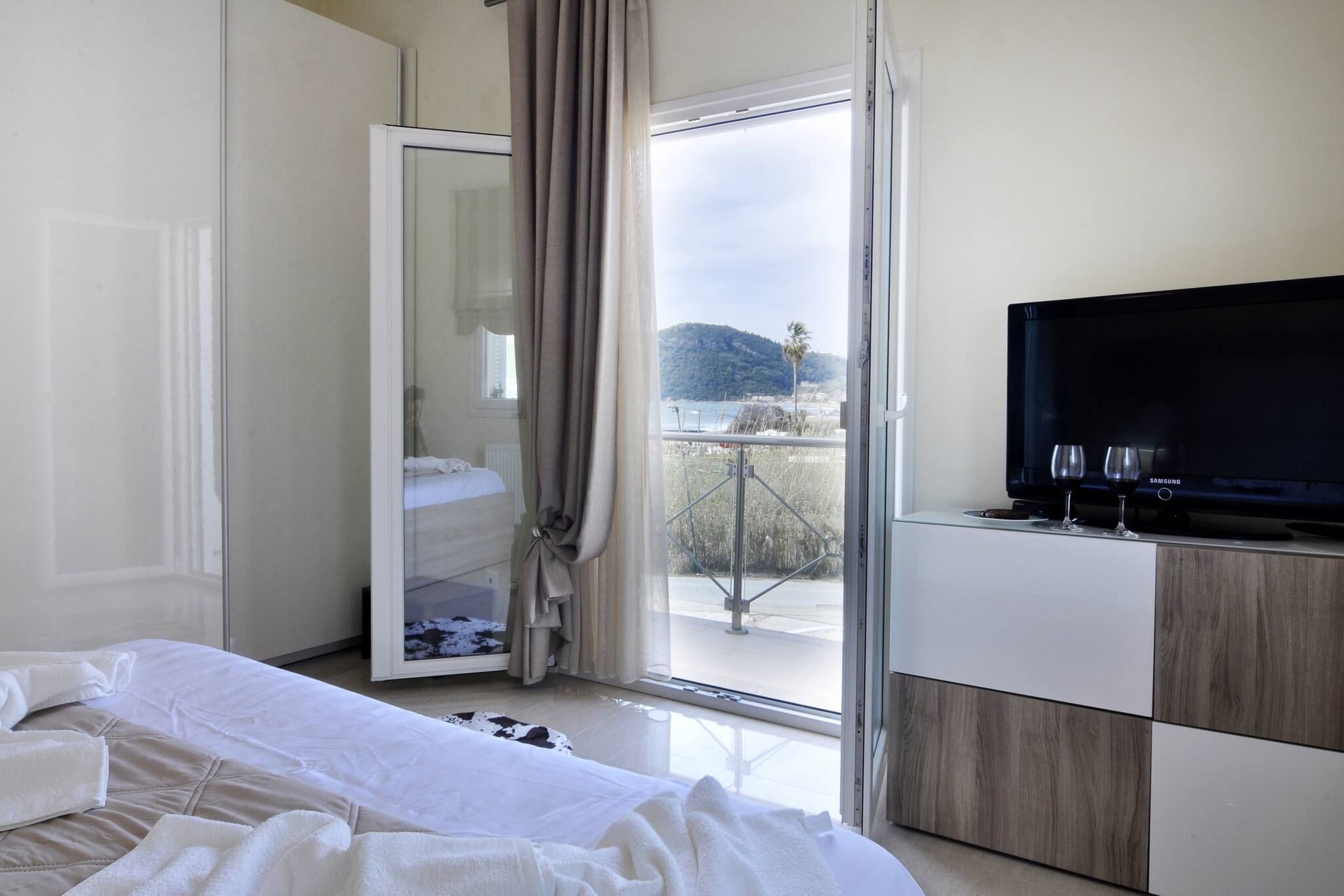 A hotel room with a bed, a TV, and a view of a beach and mountains through a sliding glass door.