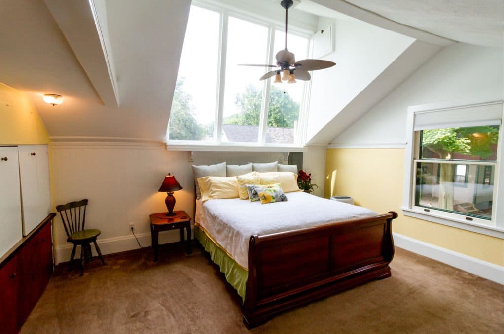 A bedroom with a sloped ceiling, a bed, a chair, a nightstand, and a window.