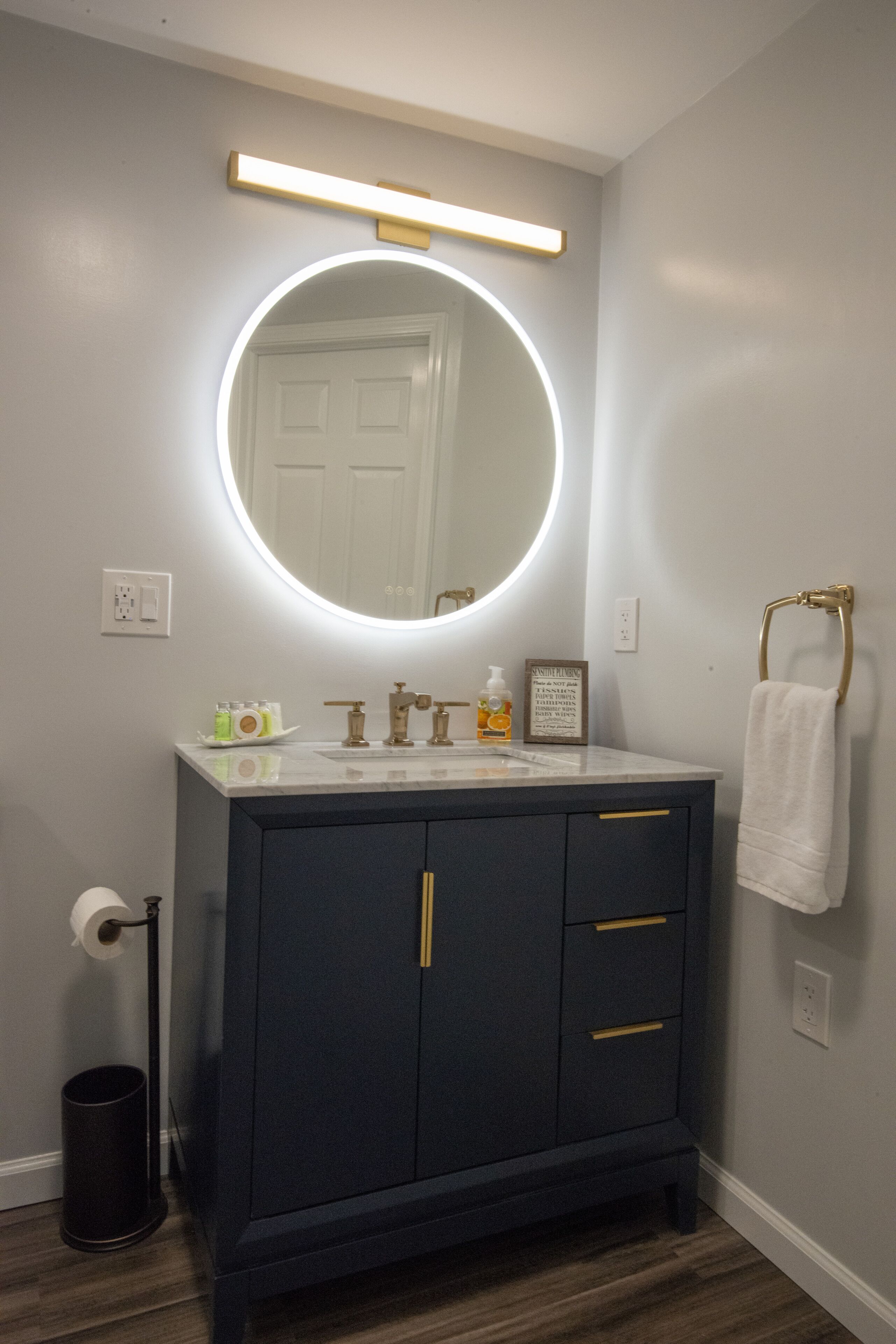 A bathroom with a dark vanity, a round mirror with a light, a towel rack, and toiletries on the counter.
