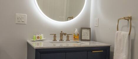 A bathroom with a dark vanity, a round mirror with a light, a towel rack, and toiletries on the counter.