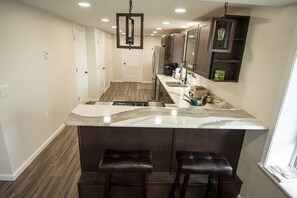 A kitchen with a marble countertop, two bar stools, wooden flooring, and dark cabinetry.