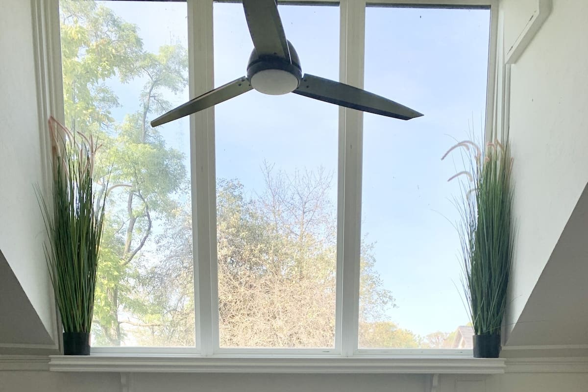 A bedroom with a bed, two potted plants, a ceiling fan, and a window with a view of trees.