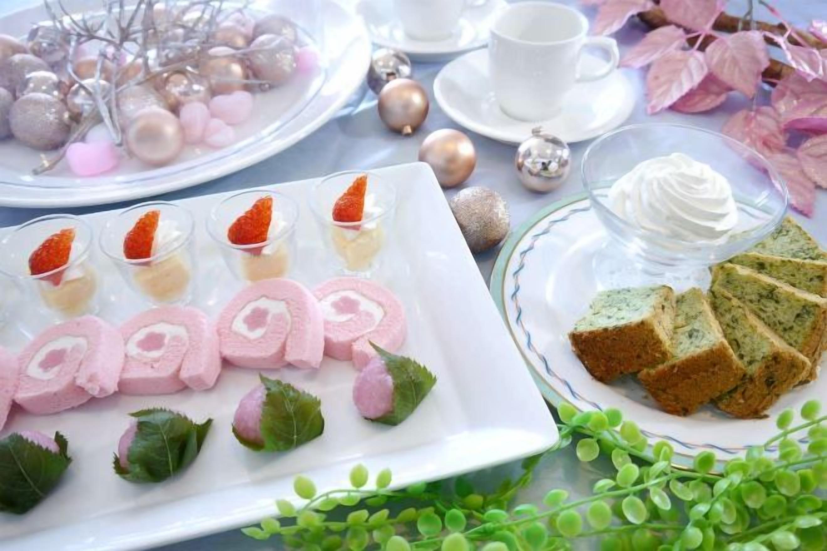 Mesa posta com diversos alimentos, incluindo um prato com pães cor-de-rosa, uma tigela de chantilly e um prato com fatias de bolo verde.