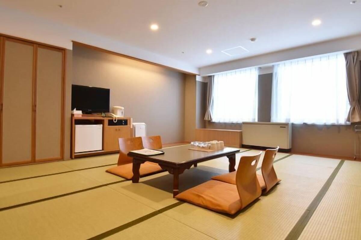 Quarto de hotel com piso de tatame, mesa de jantar com almofadas, televisão e cozinha compacta.
