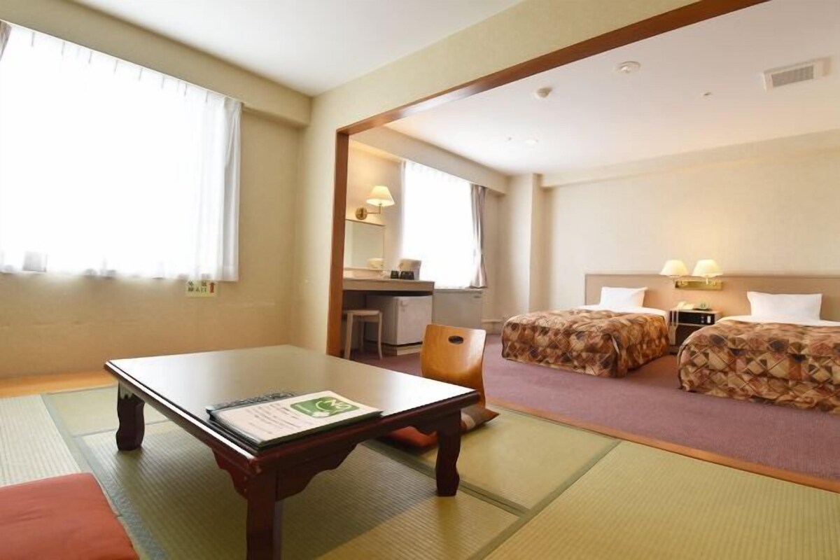 A hotel room with a tatami mat floor, a desk with a chair, two beds with floral bedding, and a small kitchenette.