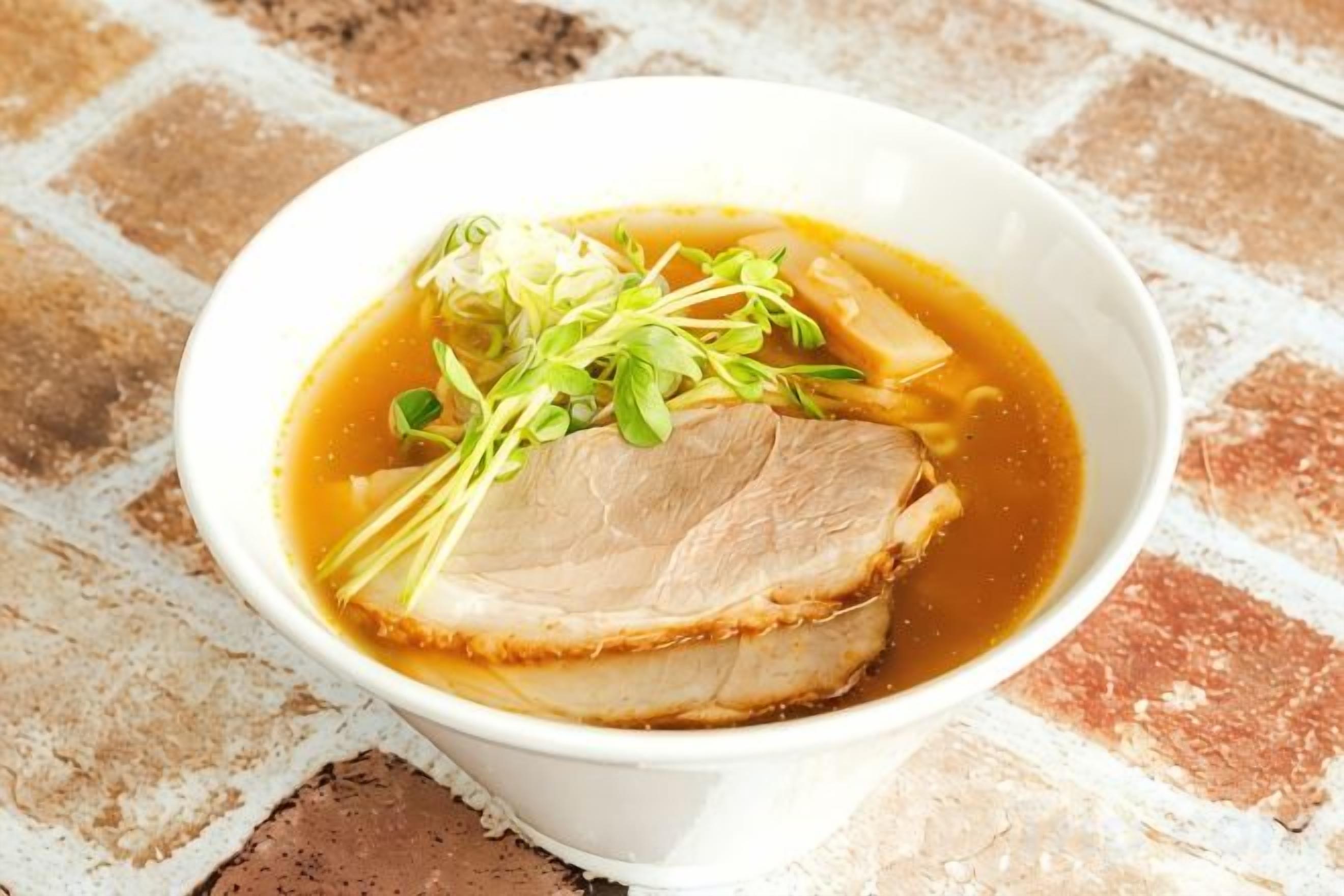 A bowl of soup with a slice of meat and greens on a brick surface.