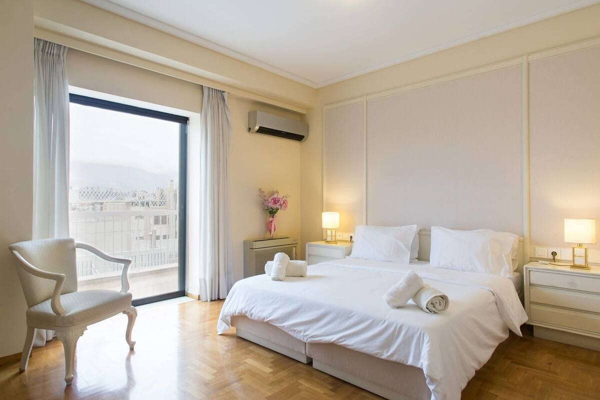 A hotel room with a large bed, two bedside tables with lamps, a chair, a window with curtains, and an air conditioning unit.