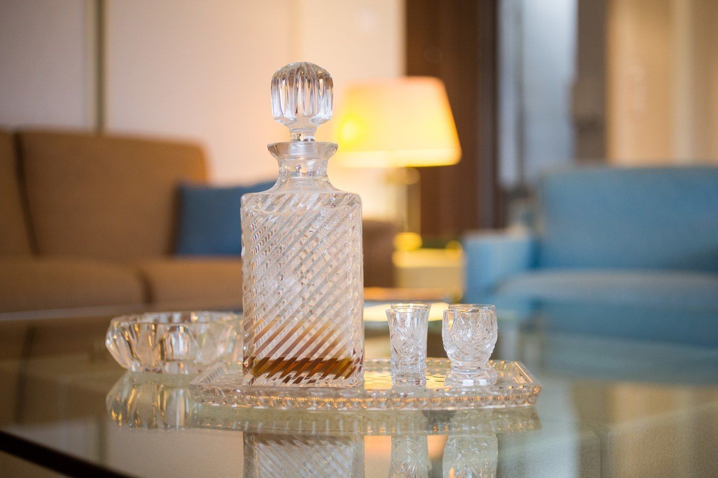 A clear glass decanter on a glass tray with two small glasses beside it, set on a reflective surface.