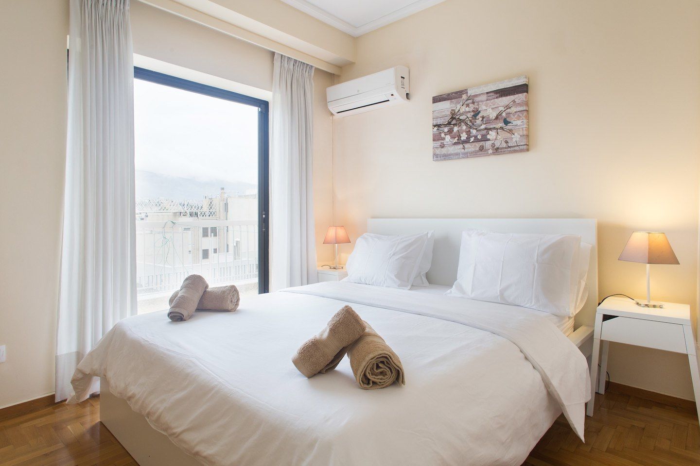 A hotel room with a large bed, white bedding, two bedside lamps, an air conditioning unit, and a city view through a large window.