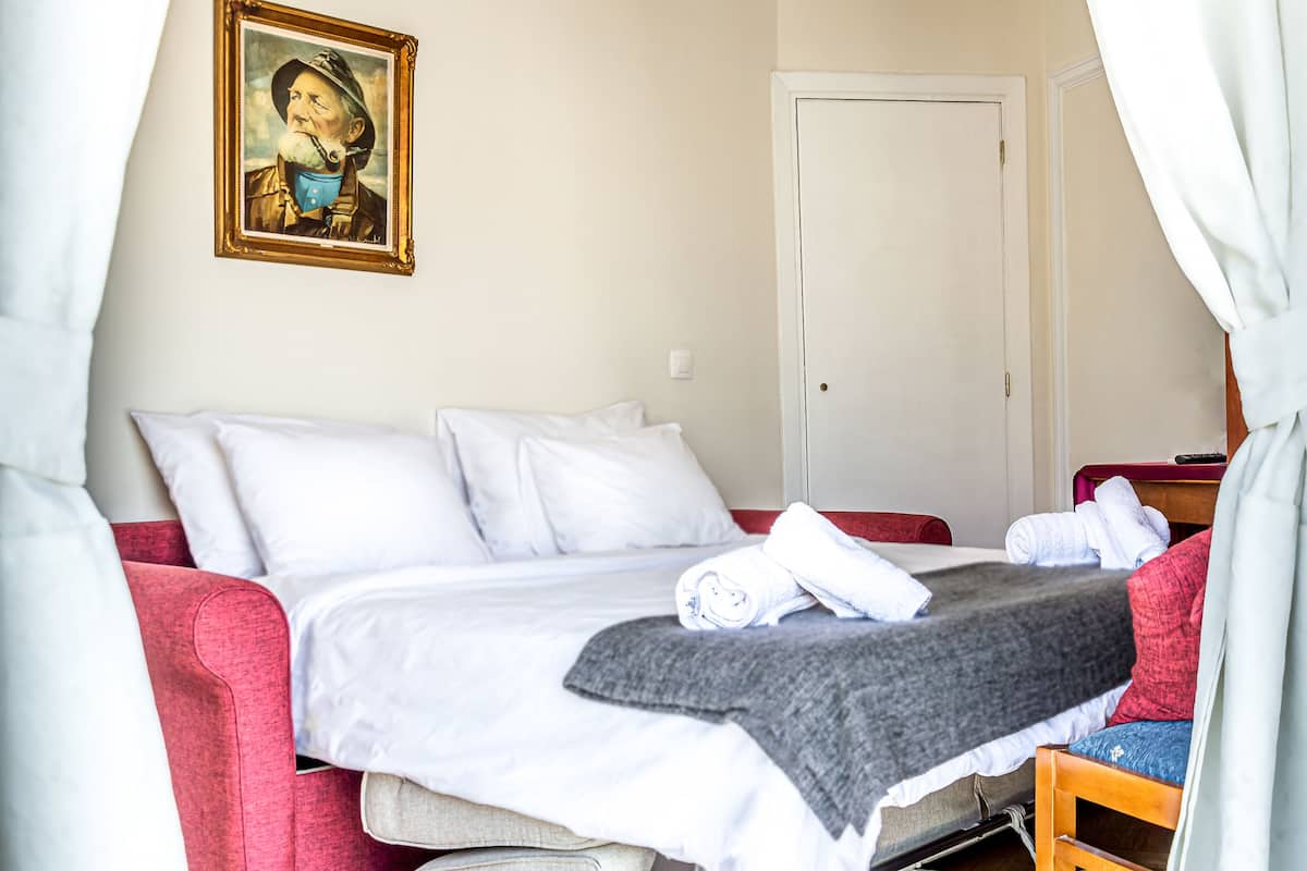 A neatly made bed with white linens and a gray blanket, a red upholstered chair, and a wooden nightstand with a lamp.