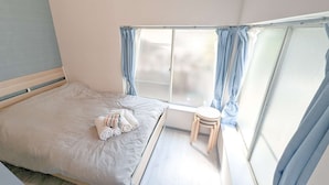 A small, single-bed room with a window, a wooden stool, and a bed with a striped blanket and rolled towels.