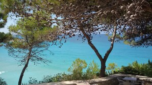 A coastal view with a clear blue sea, trees with pine cones, and a stone wall with outdoor seating. - ERATO  APARTMENT  IN ARGOSTOLI (Argostoli)