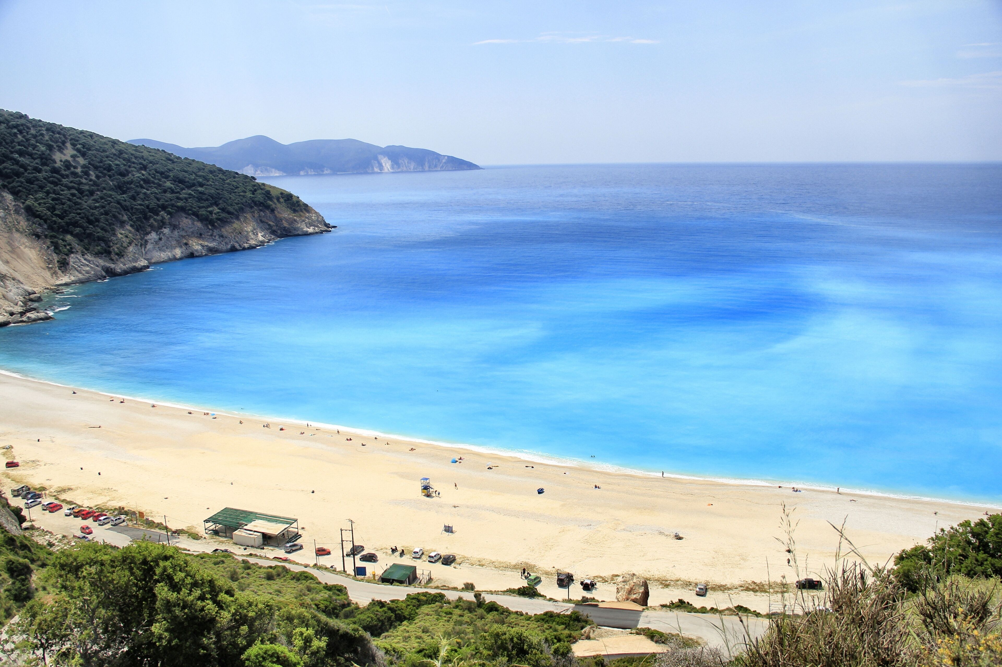 A beach with clear blue water, sandy shore, and a few buildings in the background.
