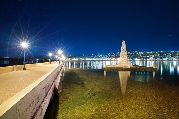 Un paseo marítimo iluminado por la noche, con un alto monumento y farolas.