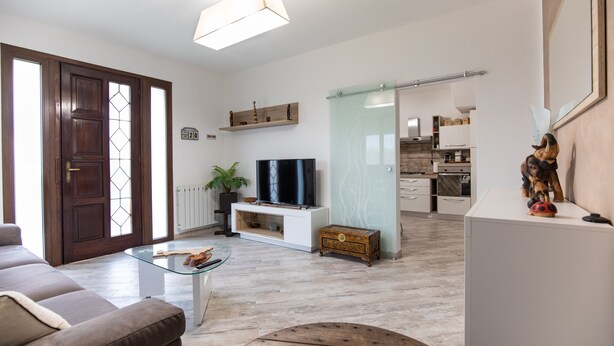 Una sala de estar con un sofá, una mesa de centro de vidrio, un televisor y una planta. Se observa un aparador de madera y una puerta de vidrio que da a la cocina.