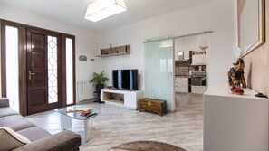 A living room with a sofa, a glass coffee table, a TV, and a plant. There is a wooden cabinet and a glass door leading to the kitchen. - Seaside villa sleeping up to 8 people with private garden and 2 parking spaces.  (Torre del Lago Puccini)