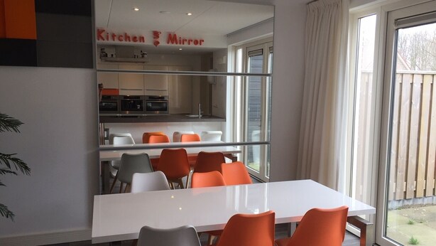 Eine moderne Küche mit einem großen Spiegel über der Arbeitsplatte, ein Essbereich mit weißem Tisch und orangefarbenen Stühlen und Blick ins Freie durch große Fenster.