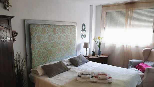 A neatly made bed with a patterned headboard, a bedside table with a lamp, and a window with blinds. - Luxury apartment in Los Alisios (Santa Cruz de Tenerife)