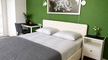 A bedroom with a bed, a desk, a chair, a nightstand, a vase with flowers, and a wall-mounted picture.