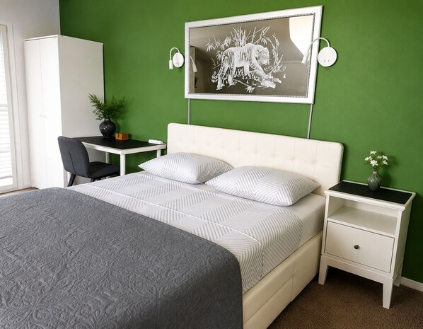 A bedroom with a bed, a desk, a chair, a nightstand, a vase with flowers, and a wall-mounted picture.