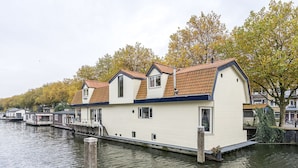 Una hilera de casas flotantes con un muelle que se adentra en una masa de agua tranquila, rodeada de árboles con follaje otoñal.