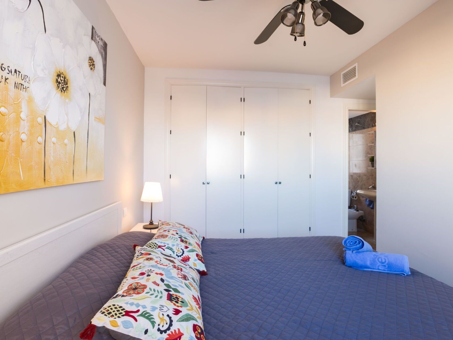 A bedroom with a bed, a ceiling fan, a wardrobe, and a bathroom visible through an open door.