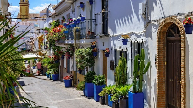 A narrow street lined with white buildings, some with balconies adorned with potted plants and flowers. - Nice apartment for 4 guests with A/C, pool, WIFI, TV and terrace (Estepona)
