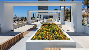 A modern architectural structure with a central garden of yellow flowers, white walls, and a clear view of the sea. - Nice apartment for 4 guests with A/C, pool, WIFI, TV and terrace (Estepona)